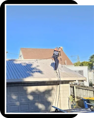Technician pressure-washing a corrugated metal roof, removing built-up moss and grime against a clear blue sky.
