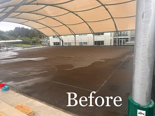 Shaded concrete court surface before pressure washing, darkened by moss and grime under tan shade sail.
