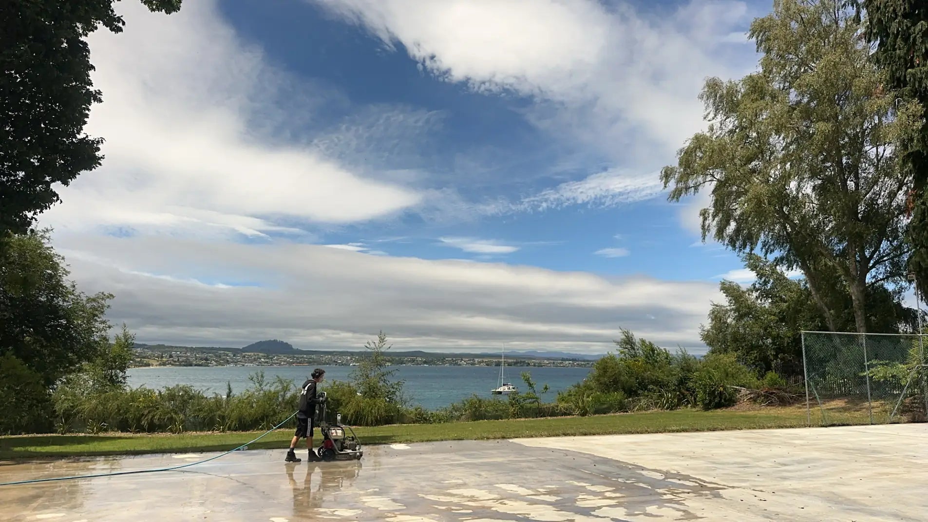 Pressure Crew technician cleaning a concrete driveway with high-pressure washer overlooking Auckland waterfront.