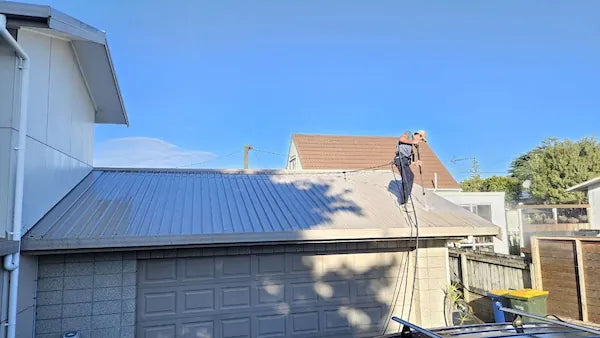 A technician stands on a corrugated metal roof under a clear blue sky, using a pressure washer to remove dirt and debris.